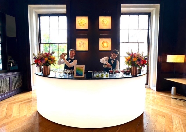 Two Bar Excellence mixologists creating cocktails behind an illuminated curved bar at a business event