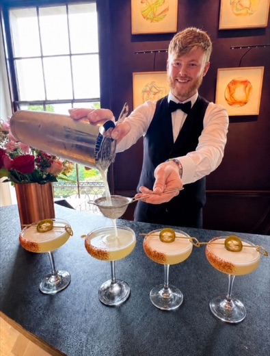 A Bar Excellence mixologist straining a fresh cocktail over coupe glasses at a live event bar