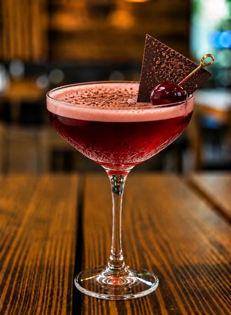 Black Forest Martini: a deep cherry-red cocktail in a coupe glass dusted with grated cacao, garnished with a dark chocolate shard and a cherry on a gold pick