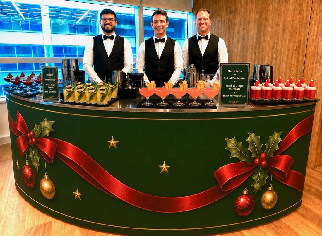 Three Bar Excellence mixologists in black waistcoats and bow ties behind a bespoke green and gold Christmas bar, with trays of Merry Berry, Peach and Ginger Margarita, Spiced Passionstar and Black Forest Martini cocktails ready to serve at a corporate event