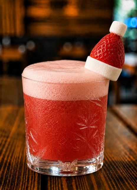 Merry Berry cocktail: a deep red drink with a pale foam top in a cut-crystal tumbler, garnished with a strawberry and marshmallow Santa hat on a gold pick